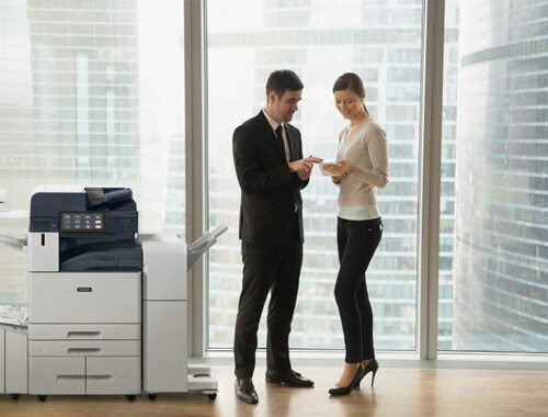 Two people in an office in business attire discussing documents stand by a Xerox® AltaLink® B8200 Series Multifunction Printer